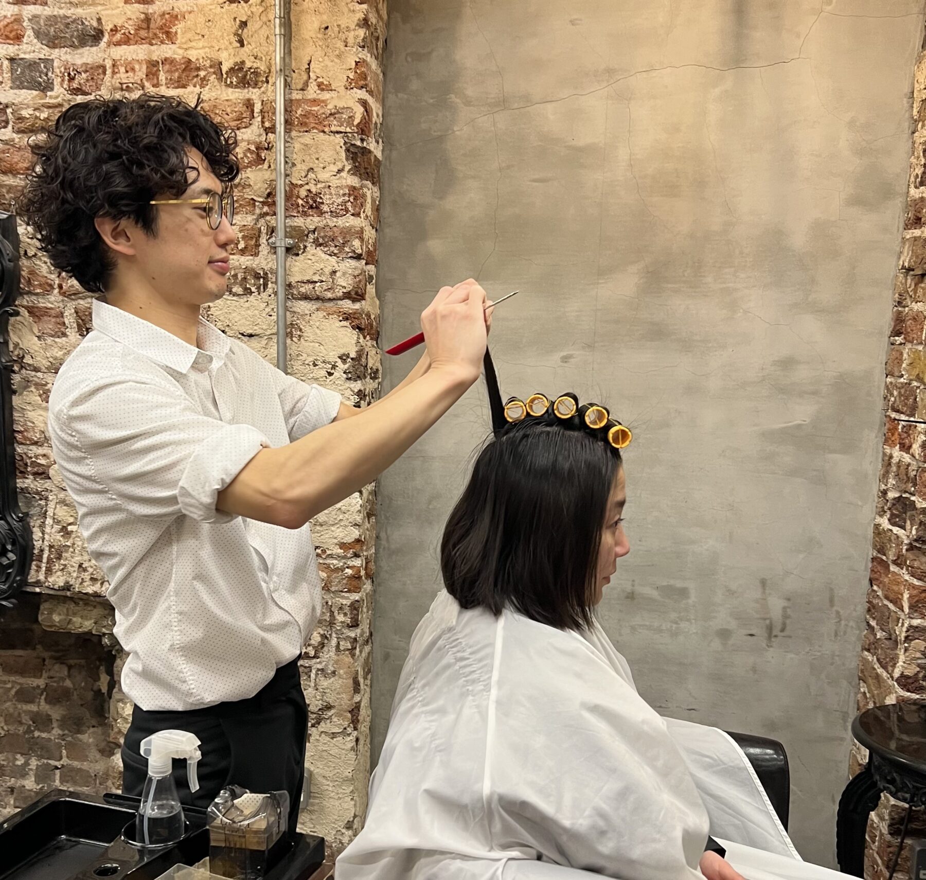“A hairstylist setting perm rods on a female client’s hair in a salon with exposed brick walls.”