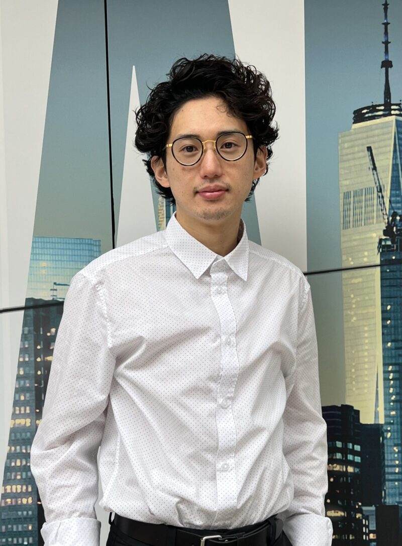 「一位穿著白襯衫、捲髮的男子,站在紐約摩天大樓的背景前。」