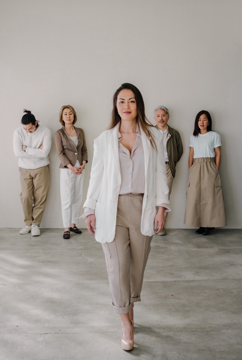 前方を歩く女性と、その後方に立つ4人の男女のグループ。全員が落ち着いた色味の服を着ている。