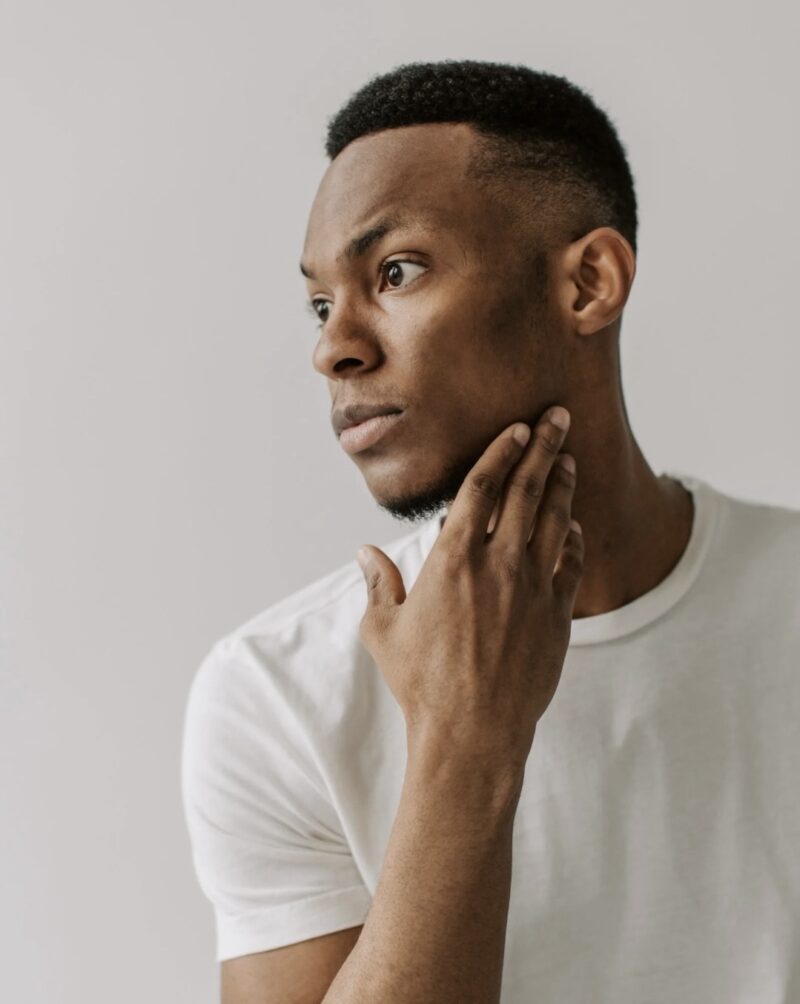 A portrait of a man in a white T-shirt gazing sideways with his hand on his chin. His hairstyle is clean and stylish.