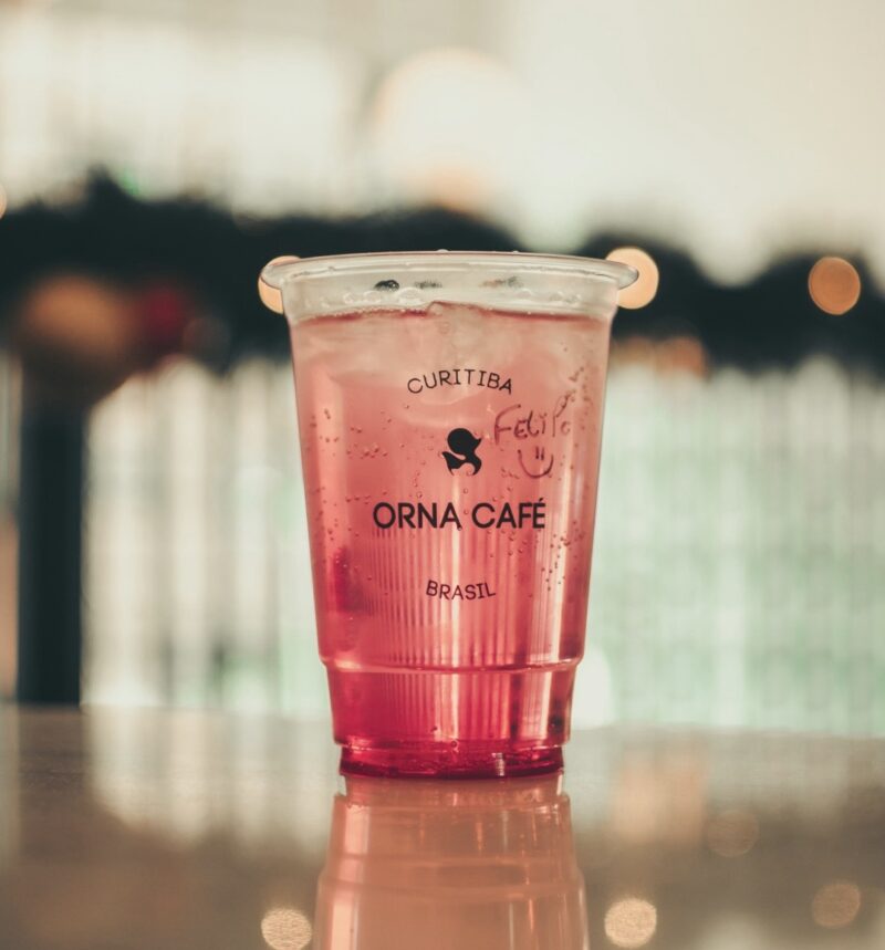 A plastic cup filled with a pink sparkling drink. The cup is labeled “ORNA CAFÉ CURITIBA, BRASIL” with a handwritten note saying “Felipe :)”.