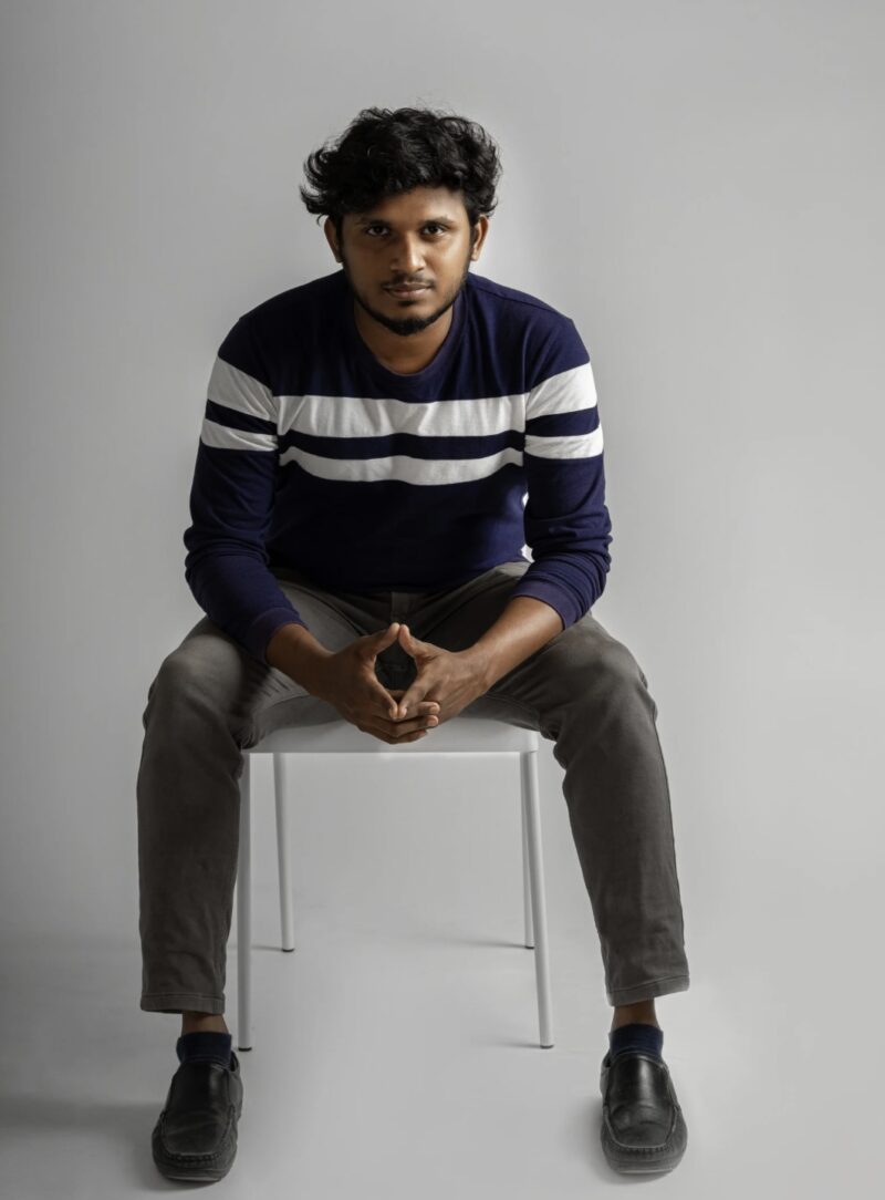 A man sitting on a white chair, looking at the camera. He is wearing a navy striped sweater and gray pants.