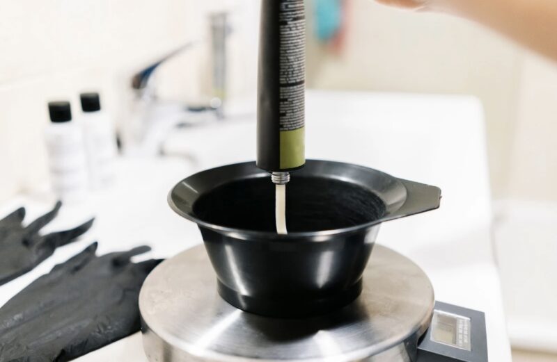Hair dye being squeezed into a mixing bowl at a salon, with black gloves and a color tube visible.