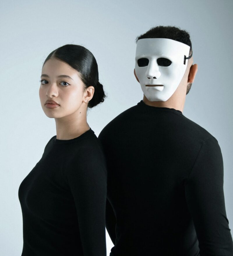 A man and a woman dressed in black stand back to back; the woman faces forward with her natural face, while the man wears a white mask and looks to the side, symbolizing contrast between individuality and appearance.