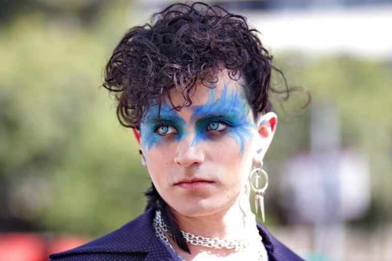 “A person with vibrant blue artistic makeup, curly dark hair, and bold accessories, showcasing a genderless street fashion look.”