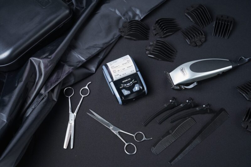 A set of barber tools neatly arranged on a black background, including scissors, clippers, combs, and hair clips.
