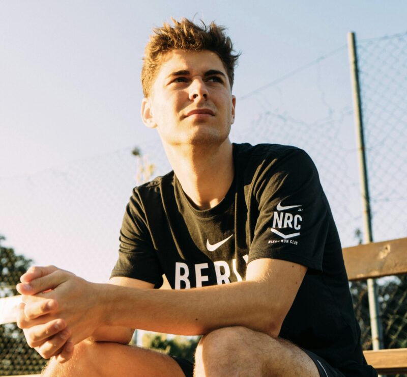 A young man with short brown hair sitting on an outdoor bench at sunset, looking into the distance. He is wearing a black Nike T-shirt and appears relaxed.