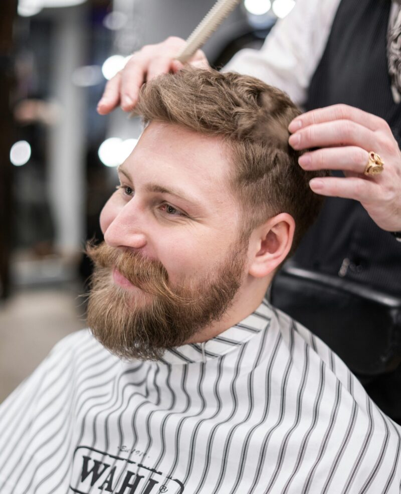A bearded man sitting in a barber chair while a barber trims the top of his hair. The man is smiling and looks relaxed during the haircut.
