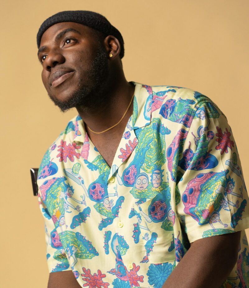 A man wearing a black knit cap and a gold necklace is looking slightly upward with a gentle smile. He is dressed in a brightly colored short-sleeve shirt featuring playful, cartoon-like illustrations. The portrait has a warm, relaxed atmosphere, set against a soft beige background.