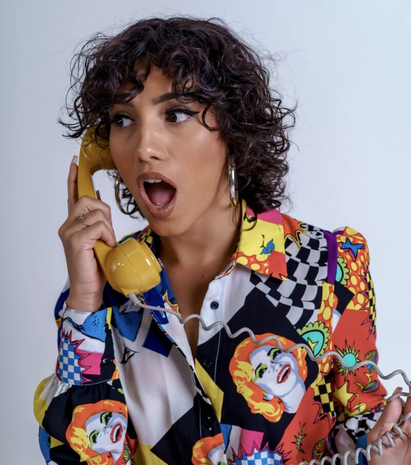 A woman wearing a colorful patterned shirt holds a yellow telephone receiver with a surprised expression. She has short curly hair and large hoop earrings.