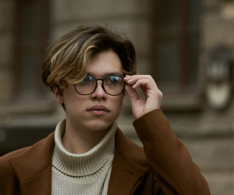 A man wearing round-framed glasses. His medium-length hairstyle has a soft, natural flow that complements the shape of his face. He is dressed in a brown coat and a turtleneck, creating a calm and refined appearance.
