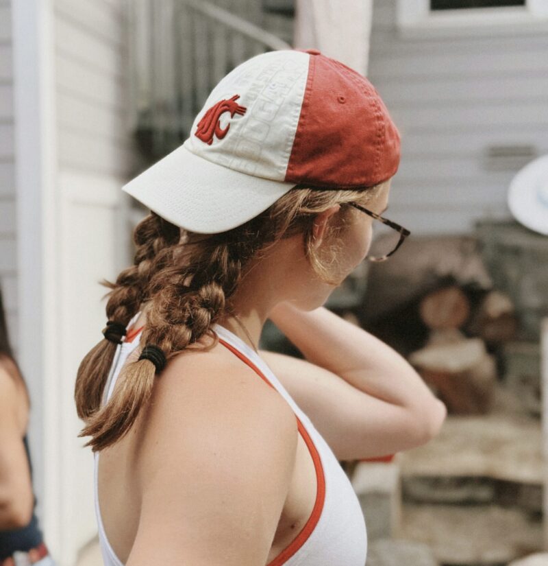 キャップをかぶり、後ろで三つ編みを2本にした女性の横後ろ姿。ナチュラルな抜け感のあるヘアスタイル。
