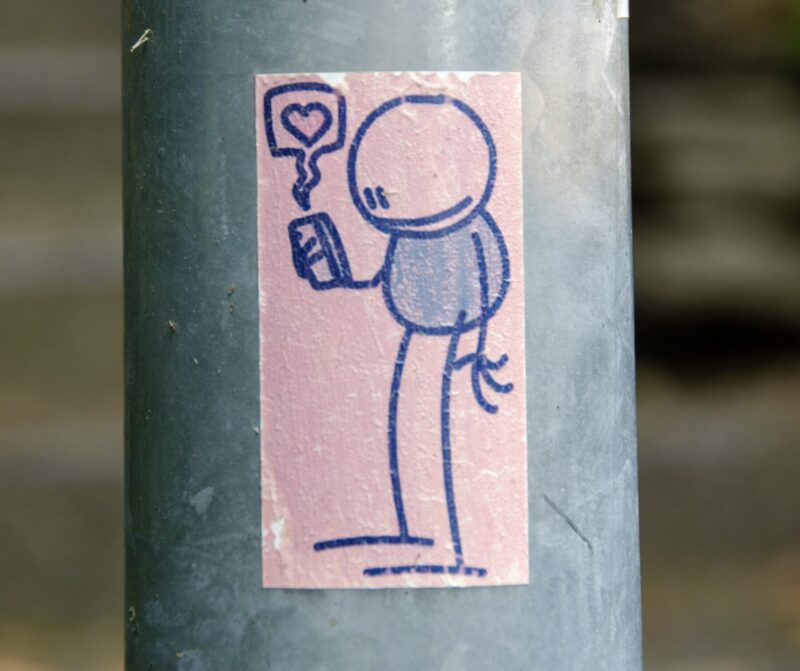 Street art of a simple character looking at a smartphone with a heart “like” icon, with a humorous and satirical element leaking from behind.